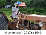 Small photo of Pelham, NH USA - July 31, 2022: A skeleton, next to an American flag, sits in the driver seat of an old rusty tractor, wearing a Let's Go Brandon t-shirt.