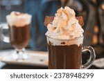 Hot chocolate with whipped cream and golden edible glitter in a transparent cup. Blurred background.
