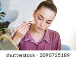 Small photo of Beautiful young woman makes blush on her face using makeup brush while sitting at home in a room at a table. Caucasian girl in a dark pink shirt does makeup on her own.