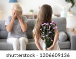 Little girl holding flowers making surprise for grandmother, congratulating her with birthday, excited granny sit with eyes closed, small grandchild prepare gift standing with bouquet in hands