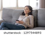 Relaxed teen girl wearing earphones listening to mobile music playing via smartphone app, calm happy young woman in headphones chilling on couch enjoying songs podcast holding cell phone at home