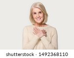 Thankful senior mature smiling woman isolated on grey studio background hold hands on chest feel appreciation and love, grateful pleased aged female look at camera thanking showing faith