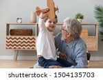 Happy two 2 generation family old grandfather and cute little child boy grandson play hold wooden toy plane lay on floor, funny small grandkid having fun with grandpa fly on airplane laughing at home