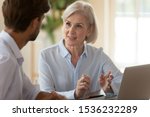 Small photo of Middle aged old businesswoman manager mentor speaking to businessman client offering insurance banking services deal teaching intern or convincing customer at business office meeting sit at table.