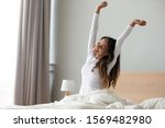 Happy woman in white nightwear sitting in bed awakened from enough and healthy sleep feels good, stretching her arms muscles after sleep and long immobility wakes up start new day with smile concept