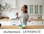 Small photo of Happy young woman holding beater microphone singing song dancing cooking alone in modern kitchen, funny lady housewife having fun listening music prepare healthy morning meal doing housework at home