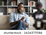 Millennial african hipster man blogger recording vlog on digital camera sit on sofa in living room, confident young guy vlogger influencer shooting social media video blog on camcorder talk at home