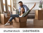 Full length overjoyed young bearded man in glasses pushing laughing wife in carton box. Energetic happy woman sitting in carboard container, having fun with smiling husband in new apartment house.
