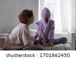 Stubborn teenage girl puts hood on head avoiding mom apology, ignoring talk with parent sitting on bed at home. Naughty teen daughter introvert hiding in hoodie showing disrespect to mother at home.