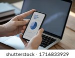 Close up businessman holding phone with graphs and diagrams on screen, analyzing startup project statistics, using smartphone, checking financial report presentation, sitting at work desk with laptop