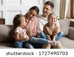 Happy young mother and father with two little daughters sitting on couch, looking at each other, family enjoying tender moment, smiling parents and preschool children having fun together