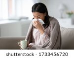 Small photo of African biracial unhealthy woman covered with warm plaid sitting on couch feels physical affliction holding mug drinking cold remedy, blow runny nose use paper tissue, seasonal flu and grippe concept