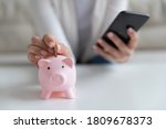 Small photo of Close up young woman putting coin in little piggy bank while managing investments, family budget or medical insurance payments in mobile banking application, personal savings expenditures concept.