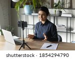 Successful Indian businesswoman recording webinar, using smartphone on tripod, sitting at desk in office, smiling young employee business coach teacher influencer shooting video for blog, speaking