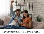 Small photo of Happy young married family couple involved in online shopping, celebrating getting gift or winning prize in giveaway, paying for goods or services with credit bank card in smartphone application.