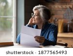Thoughtful middle aged senior woman in eyeglasses holding paper document in hands, looking in distance. Pensive smiling older mature lady feeling pleased after reading correspondence letter at home.