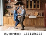 Full length energetic middle aged family couple dancing to disco music in kitchen. Happy old mature man and woman having fun, entertaining together indoors, involved in funny domestic activity.