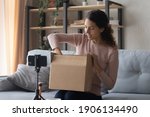Smiling pretty young female influencer unpacking big cardboard box, sitting in front of mobile phone on stabilizer, sharing internet store shopping experience with followers online or recording video.