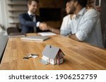 Small photo of Focus on close up keys bunch and cottage house toy model on wooden table. Smiling young man broker realtor real estate agent shake hands of happy black couple clients homeowners on blurred background