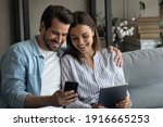 Small photo of Millennial couple in love confident internet users sit on couch hold phone tablet exchange data share information between devices. Young husband wife send receive files app from cell to pad using wifi