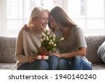 Small photo of Loving young adult female child congratulate excited elderly mother with birthday anniversary at home. Smiling caring grownup millennial daughter present gift flowers to old mom on women s day.