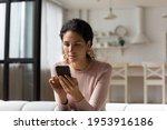 Thoughtful latin woman look at cell screen ponder on web store sales proposition reflect over answer on business offer. Interested young lady mobile phone user sit on sofa download new app for testing