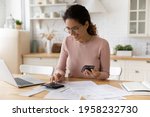 Young latina female work with financial papers at home count on calculator before paying taxes receipts online by phone. Millennial woman planning budget glad to find chance for economy saving money