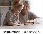 Small photo of Indoor activity. Smiling elderly female babysitter engaged in logic game with little preteen girl make move on tic tac toe or draughts board. Old granny teach grandchild to think plan in strategy game