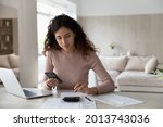Young Hispanic woman sit at desk at home manage household finances paying bills calculating on machine cellphone. Millennial Latin female care of budget use smartphone for payment. Finance concept.