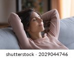 Smiling serene Hispanic woman close her eyes put hands behind head breath fresh conditioned air inside of modern living room, enjoy stress-free day off alone at home. Daydreams, peace of mind concept