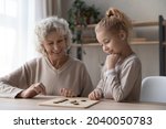 Small photo of Caring middle aged old grandmother playing draughts with adorable little child girl, sitting together at table. Happy two female generations family enjoying interesting wooden board game at home.
