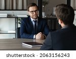 Small photo of Smart experienced young businessman job seeker in formal attire sit at desk in potential employer office pass interview answer hr recruiter question. Two millennial business partners hold negotiations
