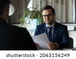 Smiling hr manager sit at desk opposite male candidate on vacant post study cv hold job interview. Young man recruiter in formal attire glasses ask applicant questions about professional experience