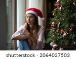 Frustrated Xmas young woman getting bored at Christmas tree, sitting at window at home, thinking over problems, feeling lonely, sad, depressed, suffering from apathy, loneliness on holiday time