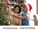 Small photo of Happy small adorable african american child girl decorating Christmas tree with happy young mother, putting toys on branches, enjoying preparing for New Year celebration at home, miracle time concept