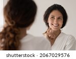 Beautiful Hispanic woman wear white bathrobe admire face view looking in mirror while standing in bathroom enjoy facial perfectness after skin procedure, applied cream, treatment for skincare beauty