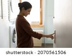 Small photo of Young Indian ethnicity female push button turns off on light inside modern domestic laundry room, adjusting thermostat for controlling air conditioner and heater inside house. Climate control concept