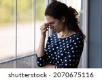 Small photo of It hurts. Exhausted young woman stand alone by window at home grimace of sudden pain headache attack touch forehead. Stressed latin female try to remember recall in memory important thing. Copy space