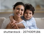 Small photo of Cheerful cute Indian preschooler kid embracing happy mother on couch, looking at camera, smiling. Young mom and kid hugging with love, affection, tenderness. Head shot home portrait