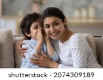 Small photo of Cute little son telling secret to cheerful Indian mom, whispering in ear. Happy loving mother and kid enjoying being friendship, trust, spending leisure time together, having fun, talking at home