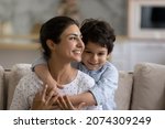 Cheerful Indian preschool kid hugging happy mom with love, gratitude, tenderness. Young mother and little son enjoying leisure time together, looking away, smiling. Family, motherhood concept