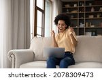 Small photo of Excited cheerful young Black woman using laptop computer on sofa at home, getting good news, feeling joy, dancing with hands, singing, laughing, making winner gesture, happy to win prize