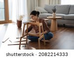 Full length smiling millennial generation indian woman sitting on warm heated floor, constructing wooden furniture in modern living room, fixing broken chair or table, improving interior at home.