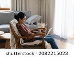 Small photo of Smiling millennial indian woman sitting on cozy armchair with laptop on laps, involved in communicating in social network, web surfing information, posting in personal blog, pending time online.