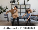 Small photo of Excited funny Black mom and curly haired daughter kid teaching dance to music in living room, enjoying activity, exercising at home, laughing. Elder and younger sisters having fun at home