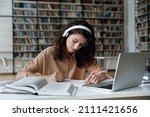 Small photo of Serious busy hardworking student girl in headphones working essay, study project in college public library, attending online learning conference, watching video lesson on internet, writing notes