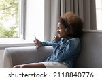 Happy gen Z Black teenage girl in big wireless headphones listening to music from player, using online media service on smartphone, making video call, talking on mobile phone