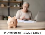Small photo of Toy pink piggy bank on work table of senior tenant, homeowner woman. Elderly lady using calculator, counting savings, taxes. Finance management, financial insurance, personal budget concept