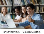 Happy millennial Jewish male student in eyeglasses showing thumbs up gesture, using computer, working on online project or preparing for exams in college library with smiling multiracial friends.