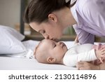 Small photo of Happy peaceful mew mom kissing cute baby lying on bed, calming infant kid for sleeping, cuddling child with love, tenderness, affection, devotion. Motherhood, parenthood concept